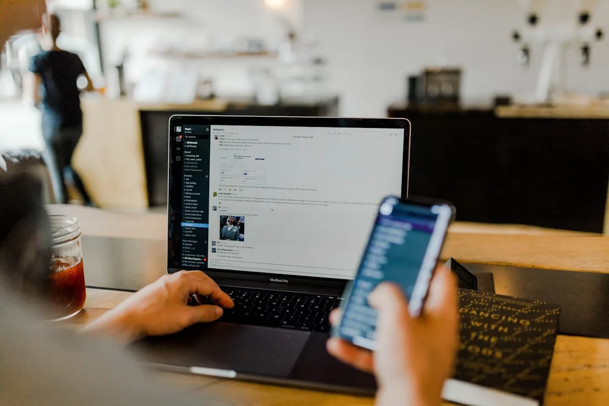 Persona trabajando en una cafetería moderna con MacBook y smartphone, representando la gestión digital de un negocio local contemporáneo