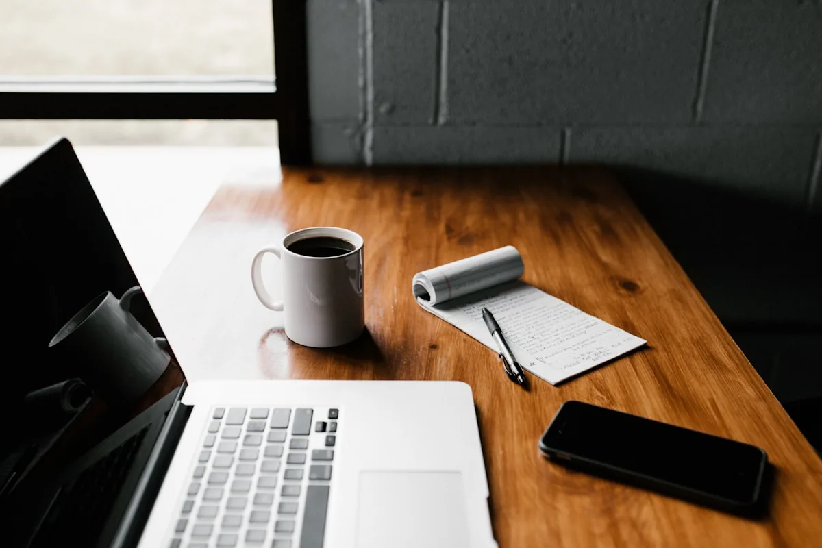 MacBook abierto junto a una taza de café, notas escritas a mano y smartphone sobre una mesa de madera, setup de trabajo para gestión de contenido digital