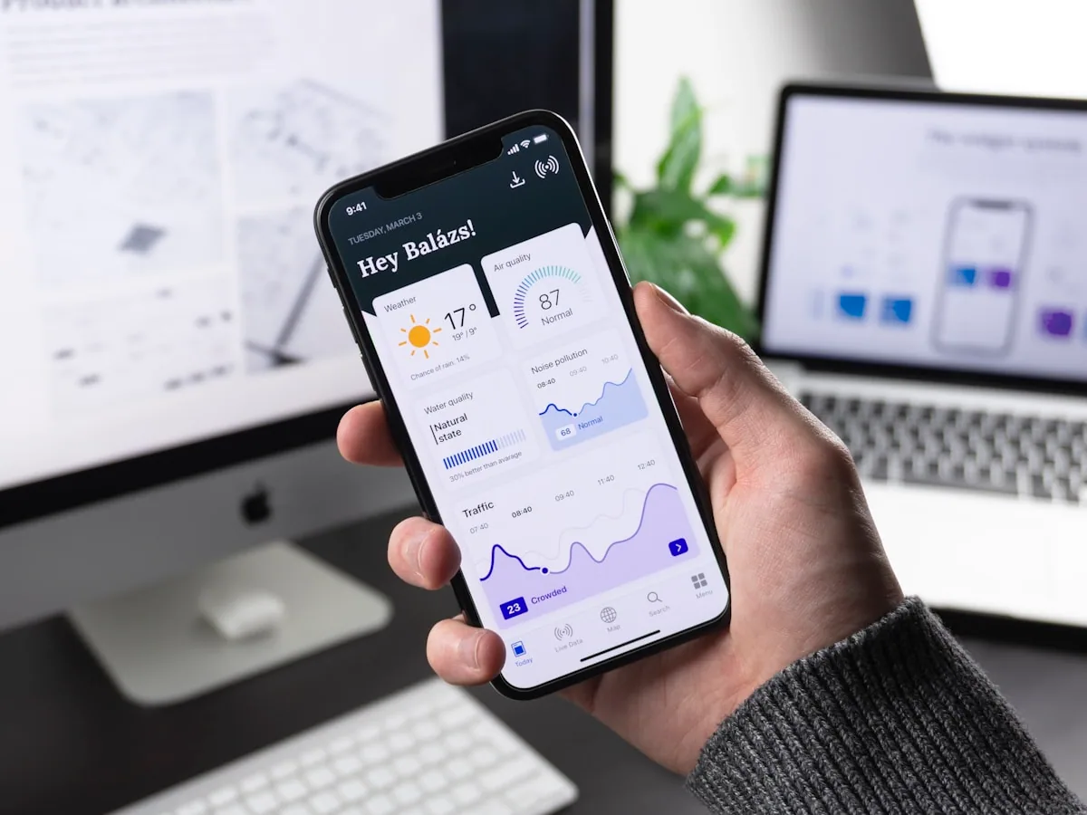 Persona sosteniendo un smartphone moderno mostrando una interfaz de app con un iMac y MacBook al fondo en una oficina