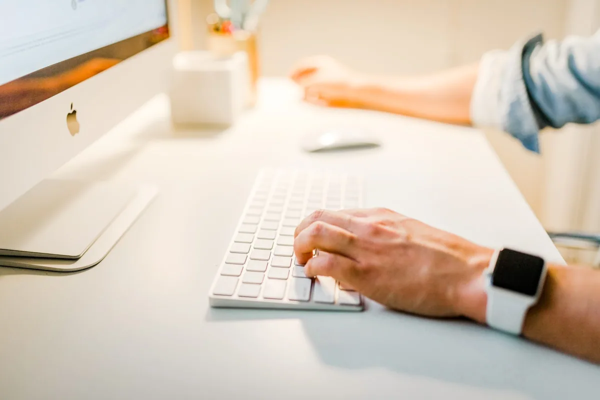 Manos escribiendo en el teclado de un iMac con Apple Watch, en un espacio de trabajo minimalista y contemporáneo